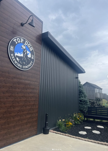 Front view of Top Dog Brewing Company sign mounted on a wooden wall beside a dark metal building, with a bench and flower bed in front.