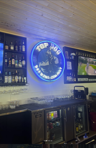 Bar interior with a blue neon Top Dog Brewing Company sign, shelves of bottles, and a row of glassware above a stainless counter.