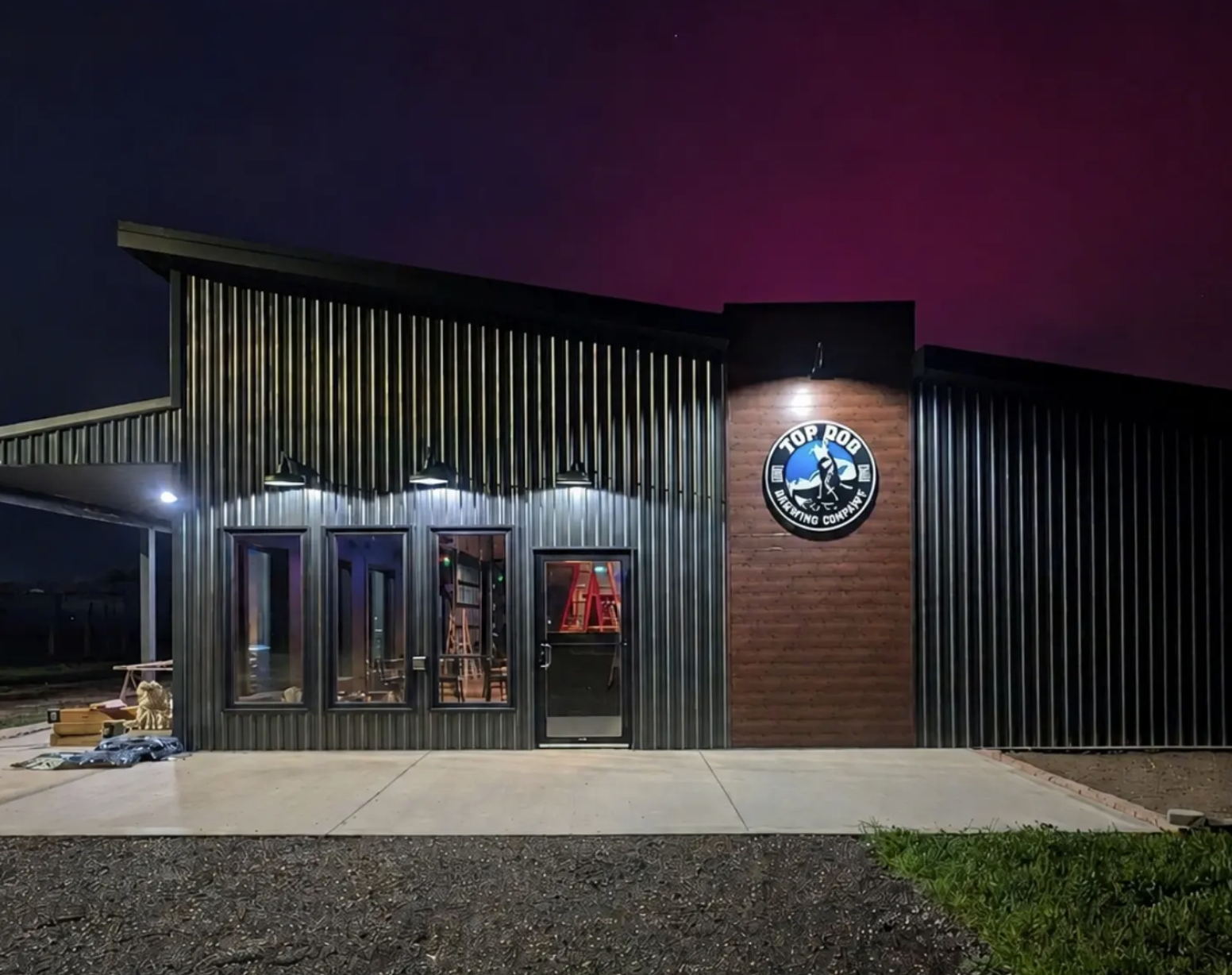 Night exterior of a modern metal-clad building with a circular logo mounted on a brick column, illuminated by exterior lights.